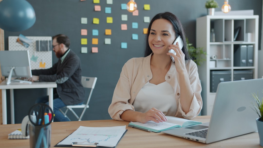 Cheerful woman is chatting on mobile phone and looking at laptop screen working in modern office. People, phoning and communication concept.